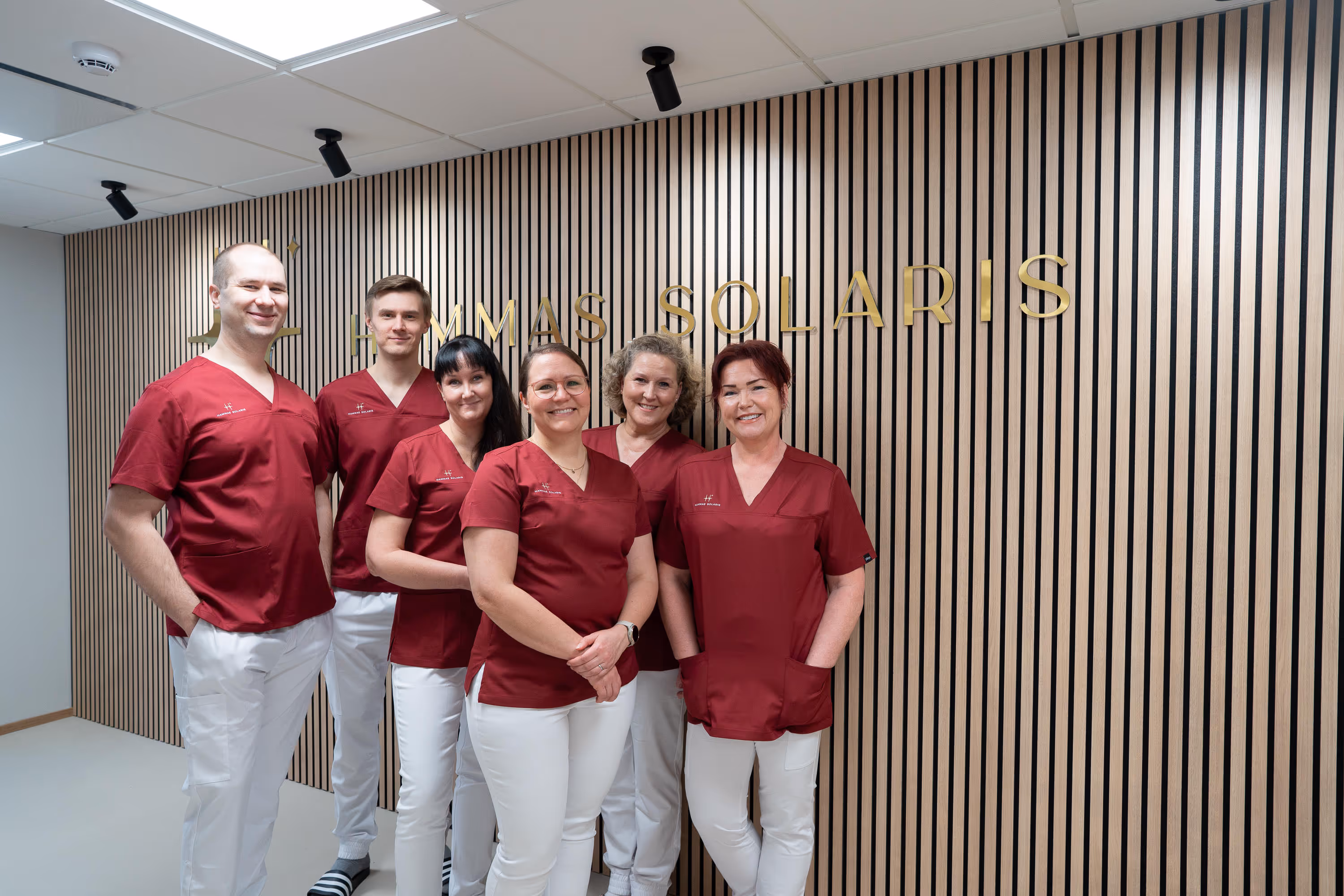 Kuusi hammaslääkäriä Naantalissa ammattilaista punaisissa työasuissa seisoo hymyillen puupaneeliseinän edessä, jossa on kultaiset kirjaimet "HAMMAS SOLARIS".
