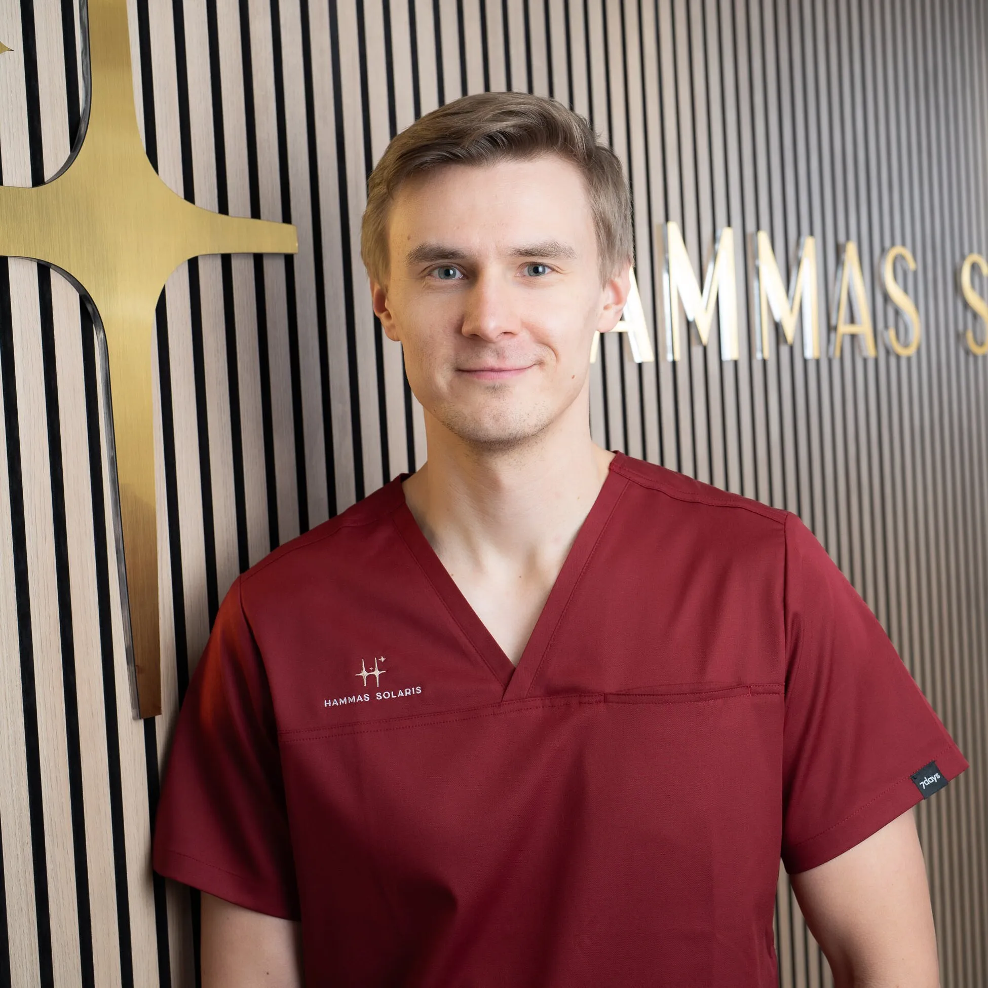 Hymyilevä mies punaisessa työasussa seisoo seinää vasten, jossa on kultainen risti ja teksti "HAMMAS SOLARIS".