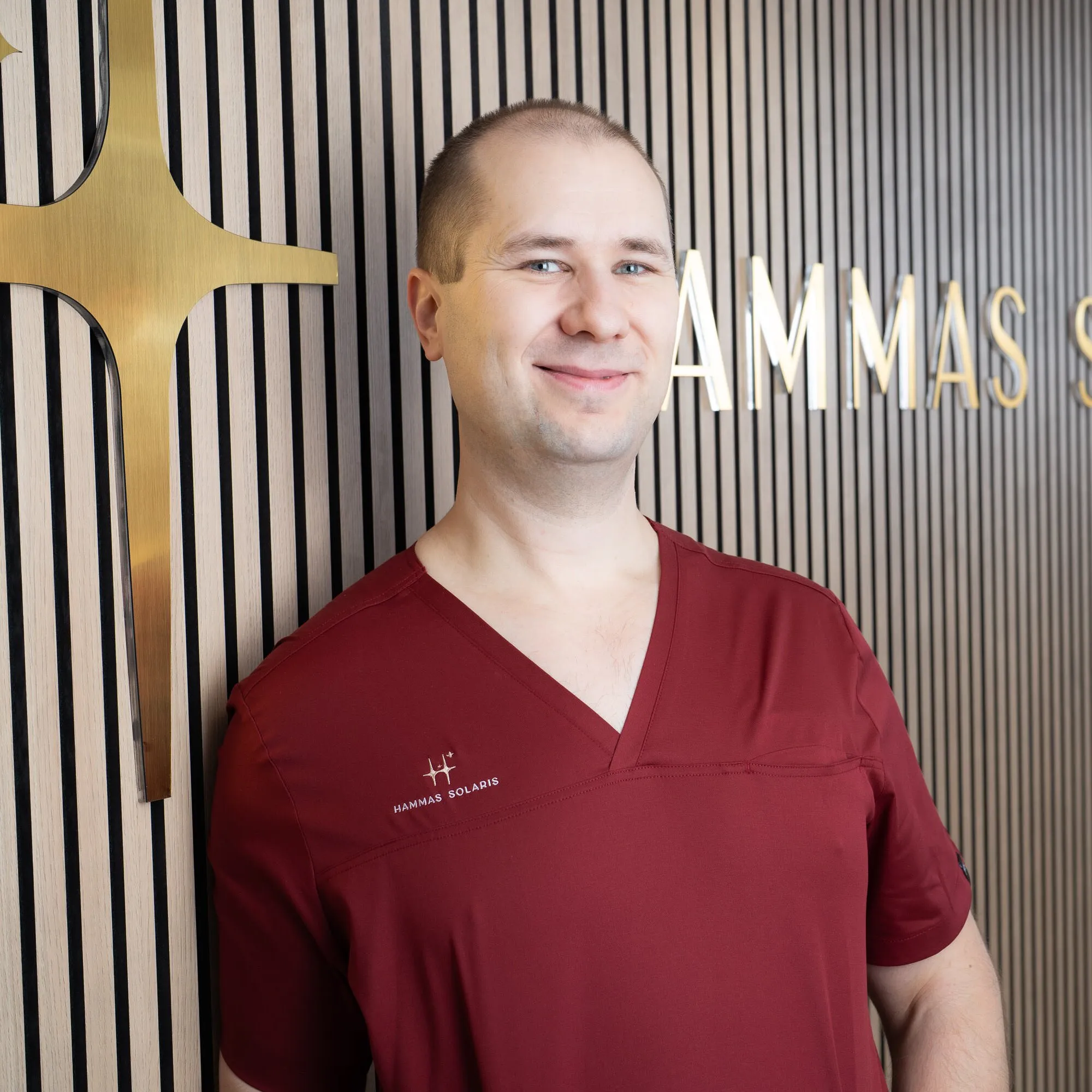 Punaisessa lääkärin univormussa oleva mies nojaa seinää vasten, jossa on kultainen risti ja teksti "HAMMAS SOLARIS". Taustalla on puupaneeli, jossa on mustia ja vaaleita raitoja.