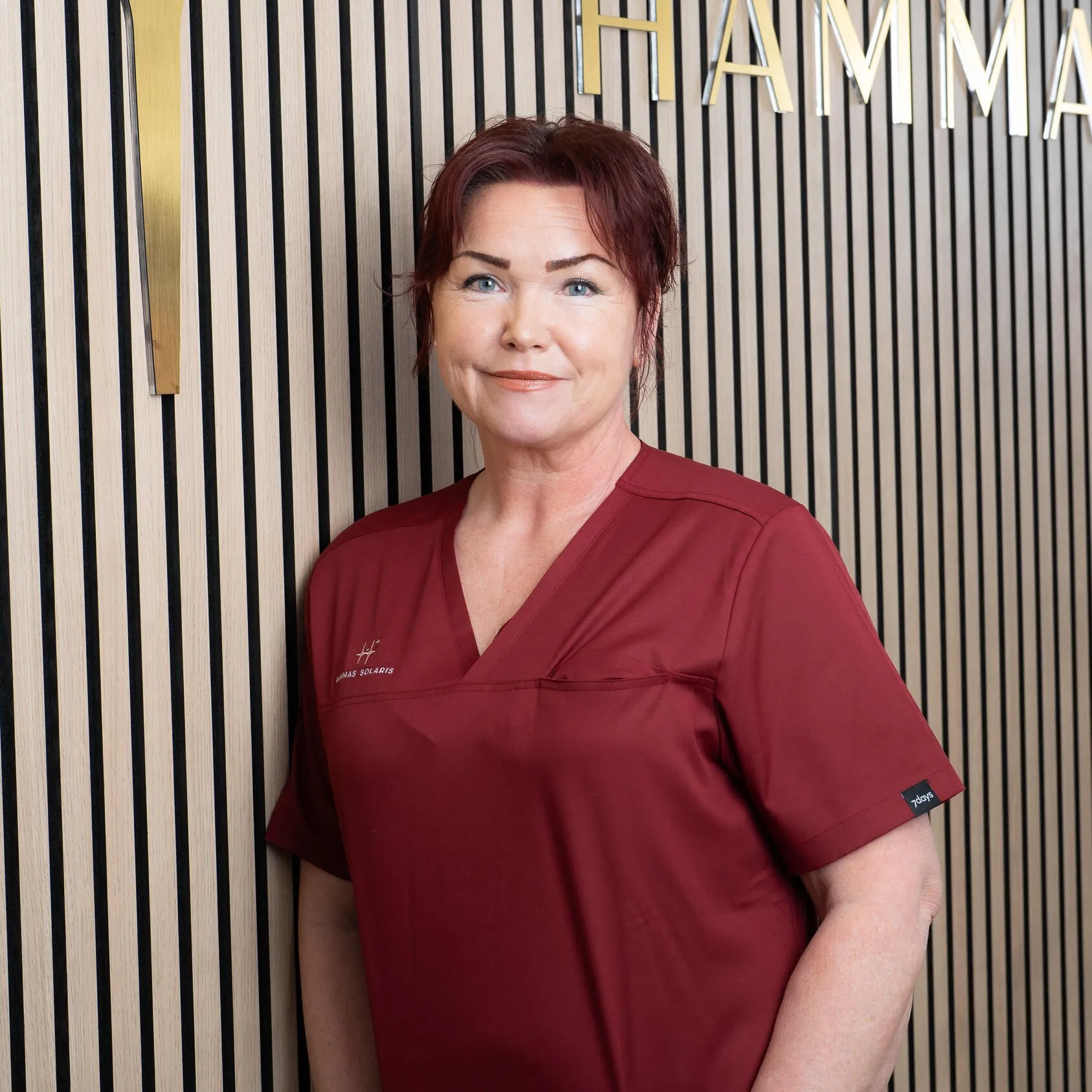 Keskellä seisova nainen punaisessa hoitopuvussa, taustalla puupaneeli ja kultaiset kirjaimet. Kirkas valaistus.