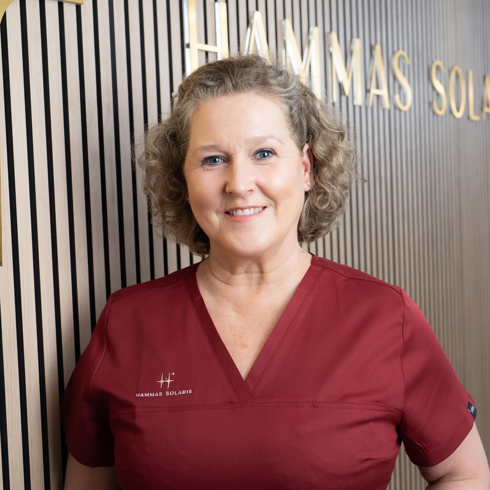 Keskellä oleva hymyilevä nainen punaisessa työvaatteessa, taustalla seinä ja teksti "HAMMAS SOLARIS".