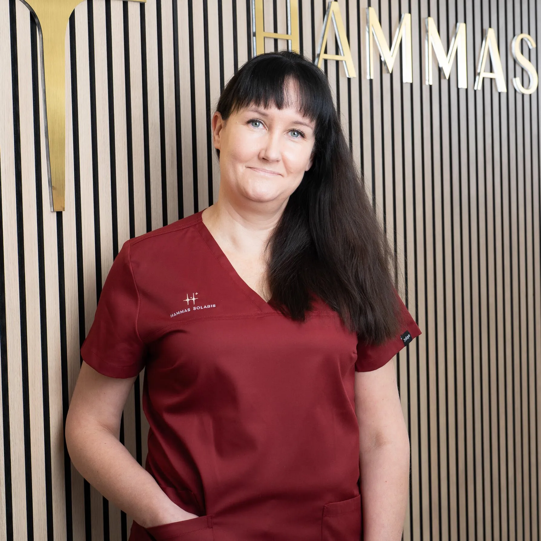 Nainen punaisessa työpaidassa seisoo seinää vasten, jossa on kultaiset kirjaimet "HAMMAS" ja pystysuorat mustavalkoiset raidat.