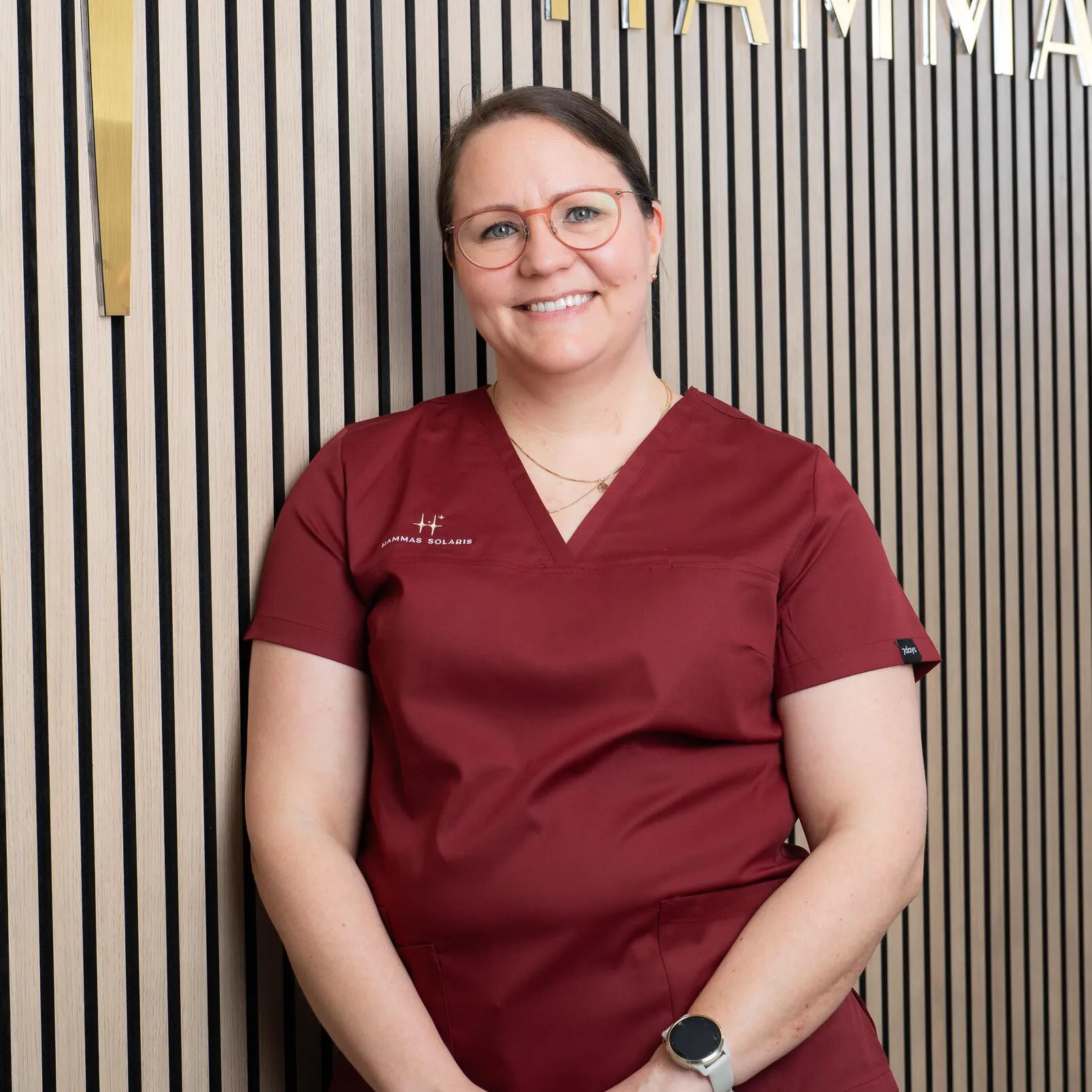 Hymyilevä nainen punaisessa asussa, taustalla puupaneeli ja kultaiset kirjaimet. Hänellä on pyöreät silmälasit ja vaaleat hiukset.